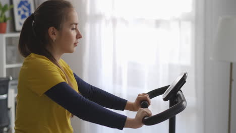 young-sporty-woman-on-stationary-bike-in-living-room-training-alone-at-weekend-sport-equipment-for-home-fitness-keeping-fit-and-losing-weight